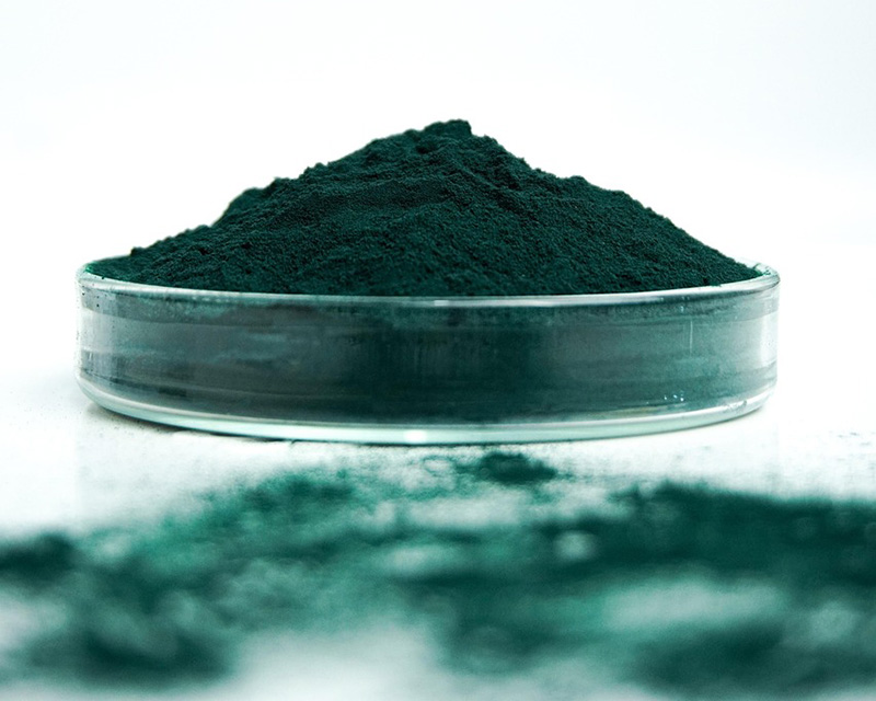 Spirulina in polvere in un contenitore circolare trasparente e sparsa sul piano bianco su cui quest'ultimo è appoggiato